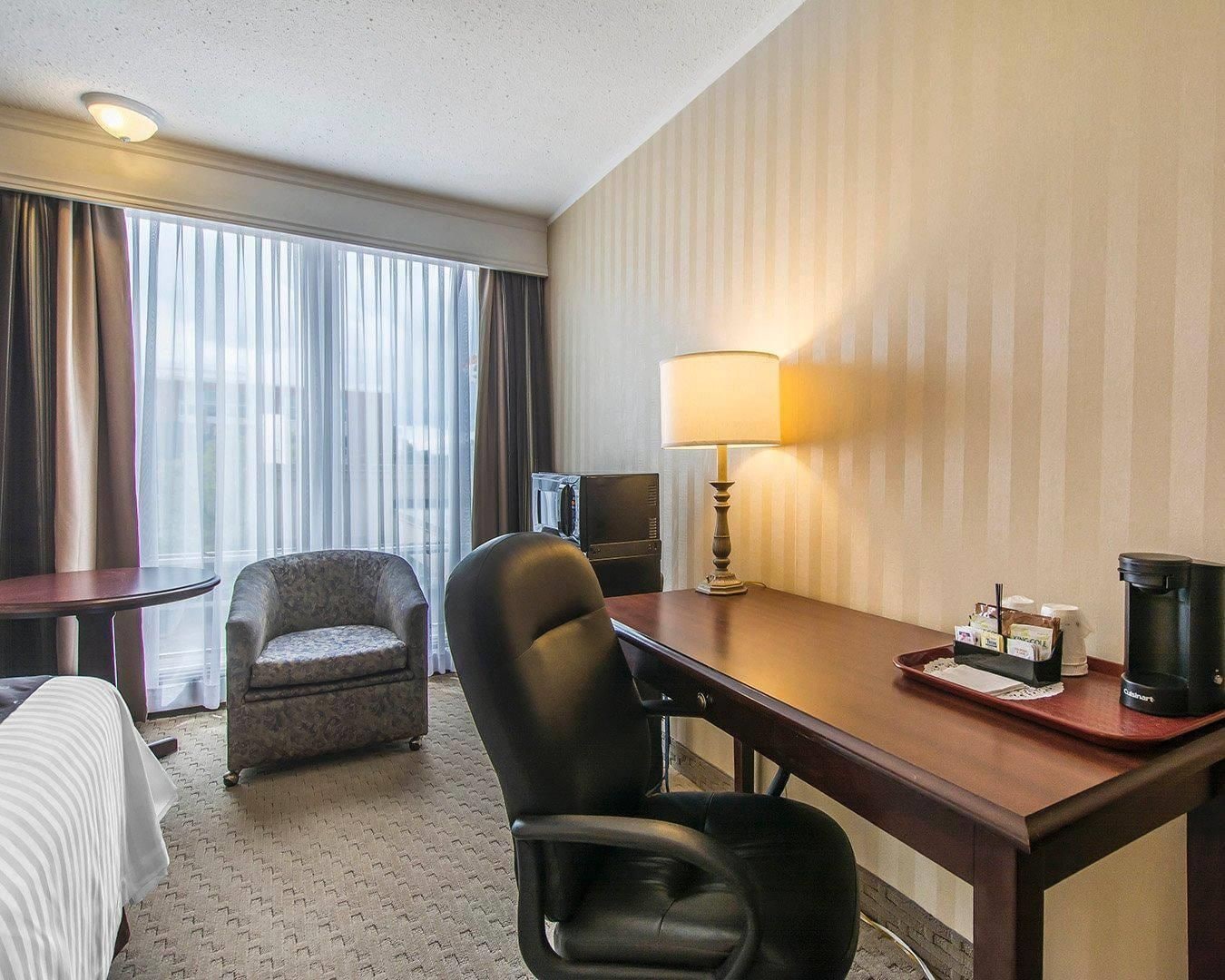 A hotel room with a bed, a desk with a lamp, a chair, a coffee maker, and a television.
