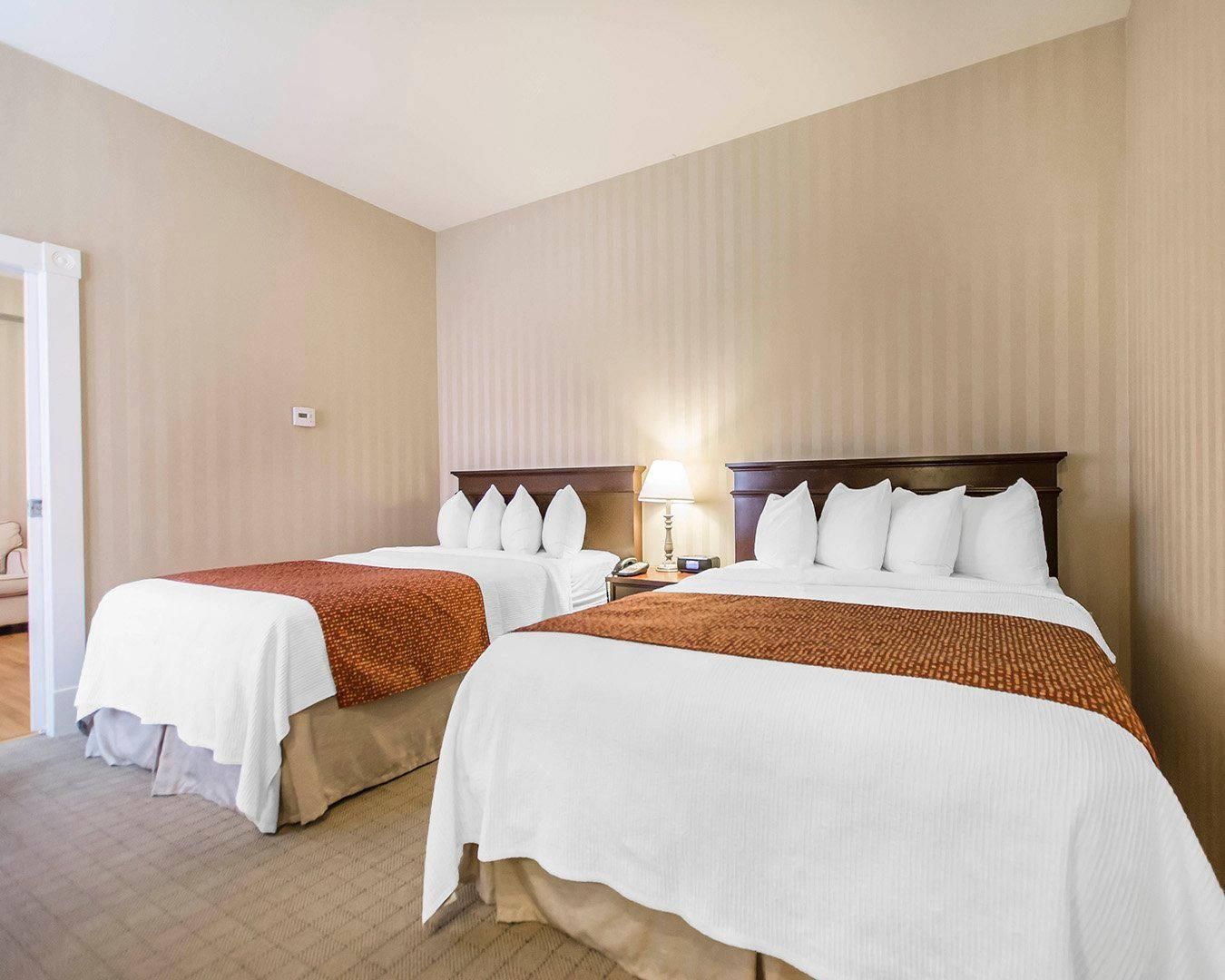 Una habitación de hotel con dos camas, una mesita de noche con lámpara y una puerta que da a un pasillo.