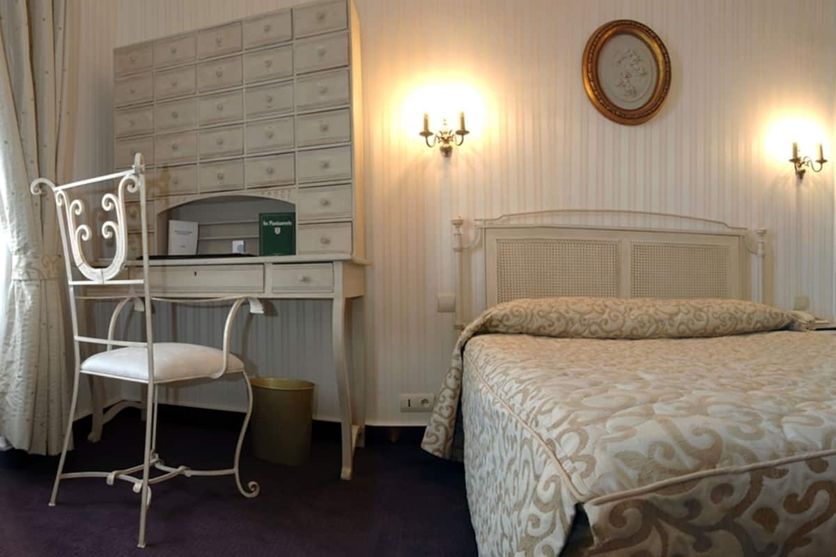 A bedroom with a bed, a desk with drawers, a chair, and wall-mounted lights.