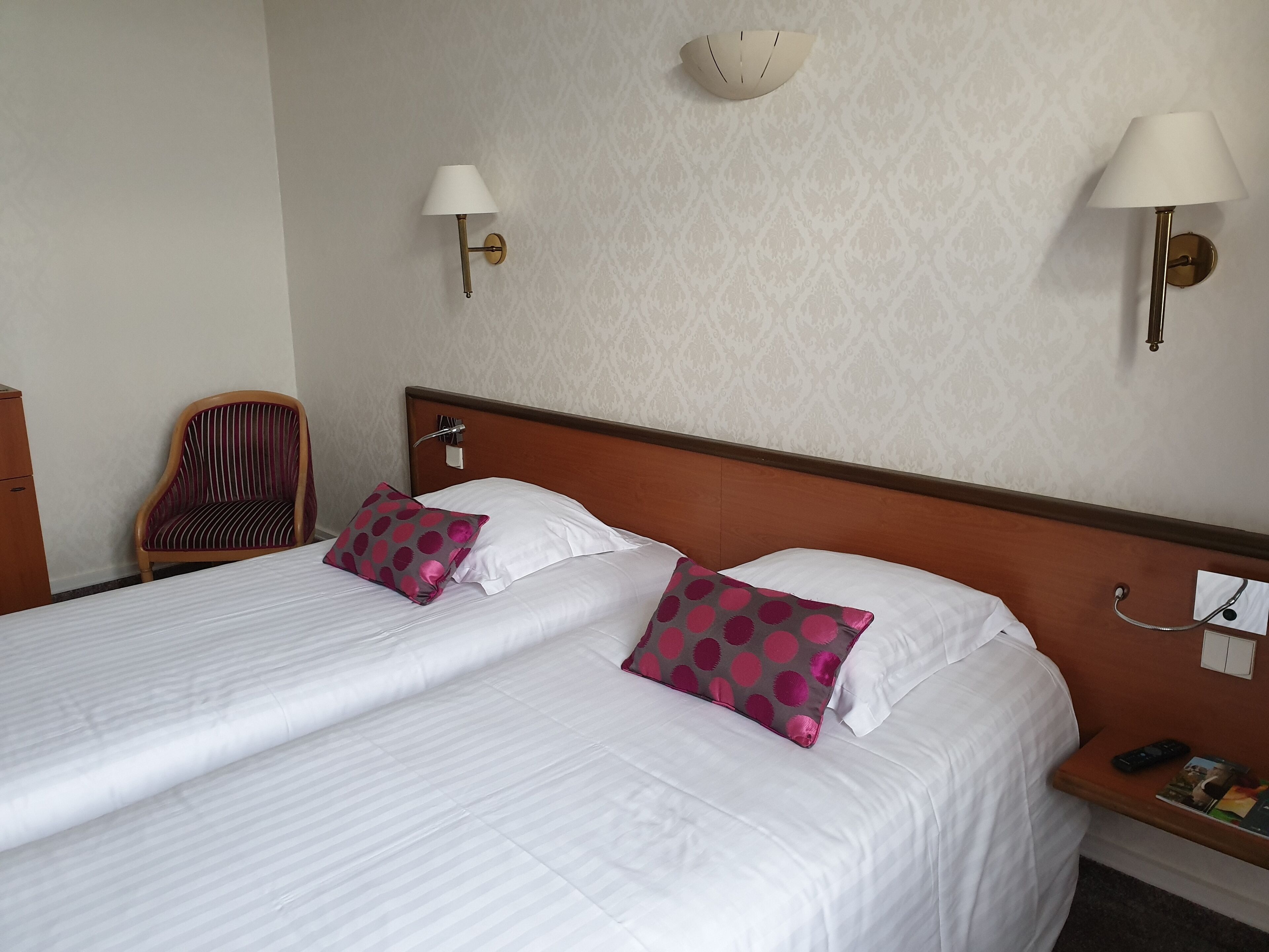 Habitación de hotel con cama doble, dos almohadas, mesita de noche de madera, una silla y dos lámparas fijadas a la pared.