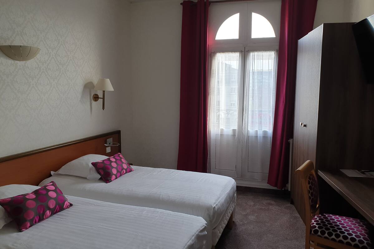A hotel room with two beds, a desk, a chair, a wardrobe, and a window with curtains.