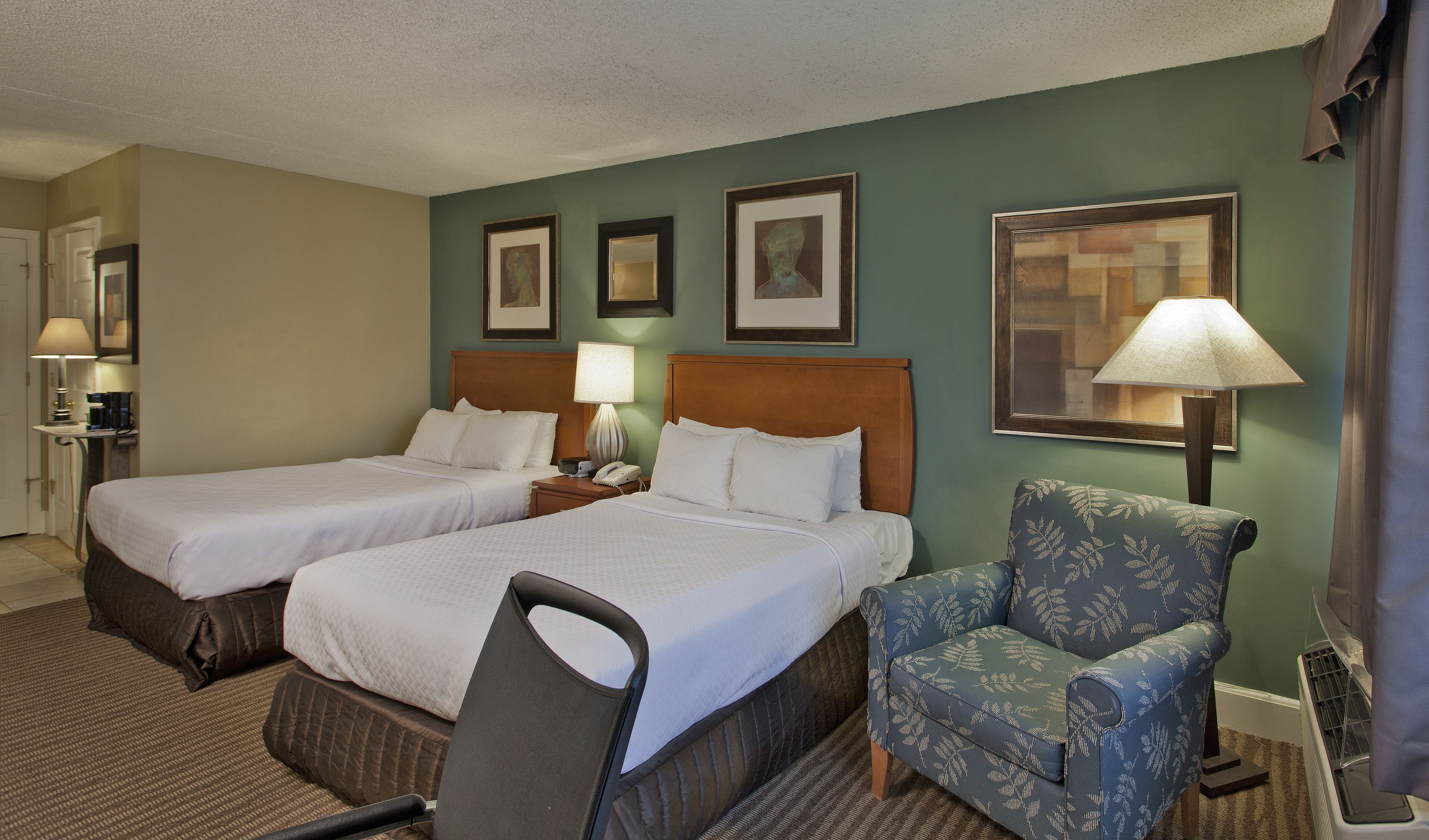 A hotel room with two beds, a chair, a desk with a lamp, and framed pictures on the wall.