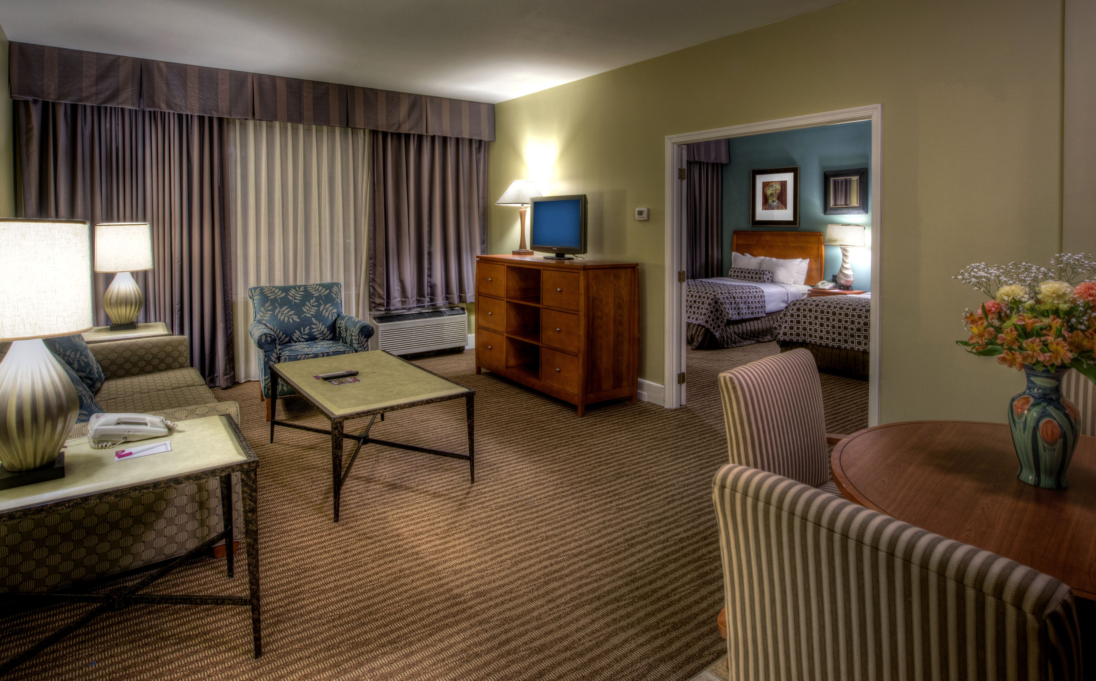 A hotel room with a sofa, armchair, a small table with a lamp, a TV on a wooden cabinet, and a bed with a headboard.