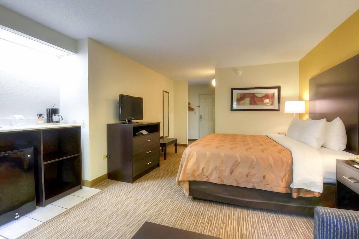 A hotel room with a bed, a desk with a television, a dresser, and a bathroom visible in the background.