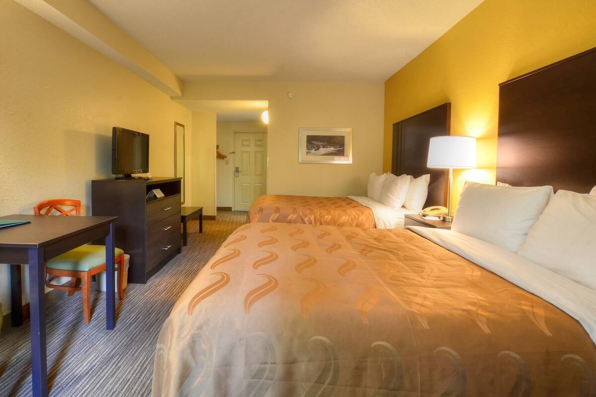 A hotel room with two beds, a desk, a television, and a lamp.