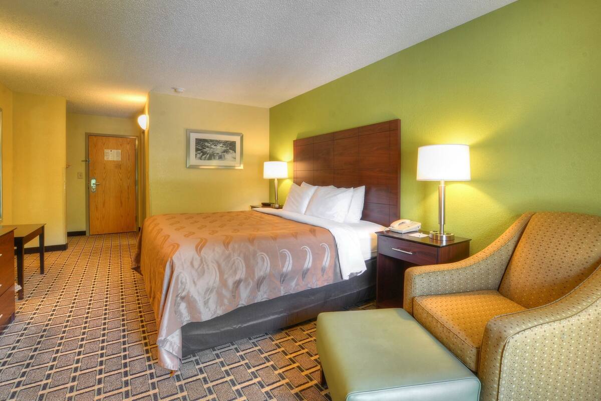 A hotel room with a bed, a chair, a desk, and a lamp.