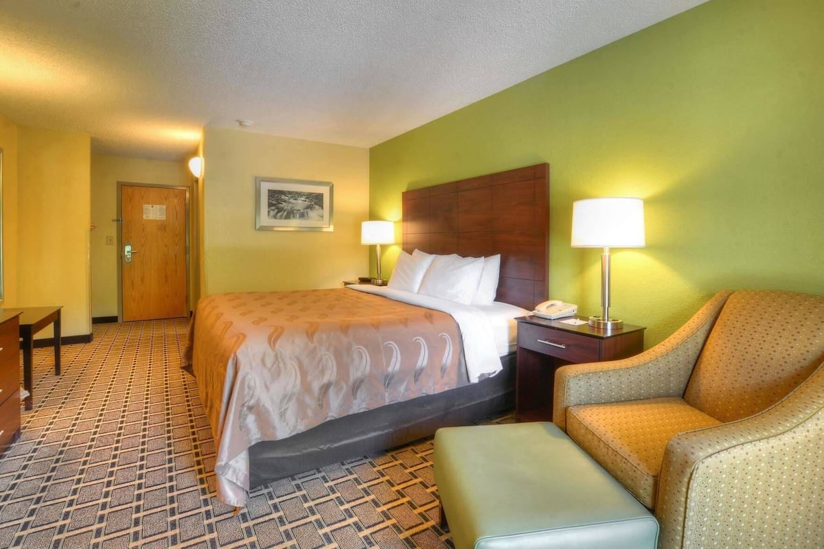 A hotel room with a bed, a chair, an ottoman, a desk, and a door.