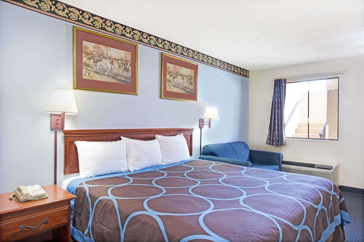 A hotel room with a large bed, two wall-mounted lamps, a telephone, a blue chair, and a window with curtains.