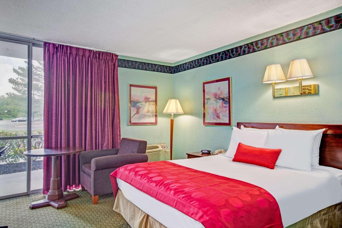 A hotel room with a bed, a chair, a table, a lamp, and a window.