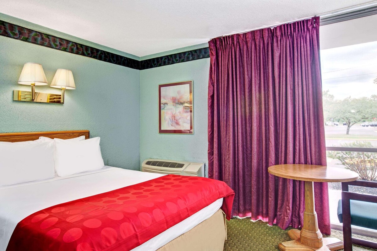 A hotel room with a bed, a table, a window with curtains, and a wall-mounted air conditioner.