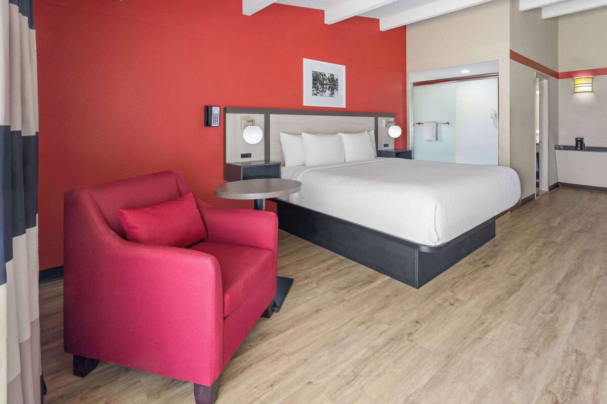 A hotel room with a bed, a red armchair, a small round table, and a nightstand with a lamp.