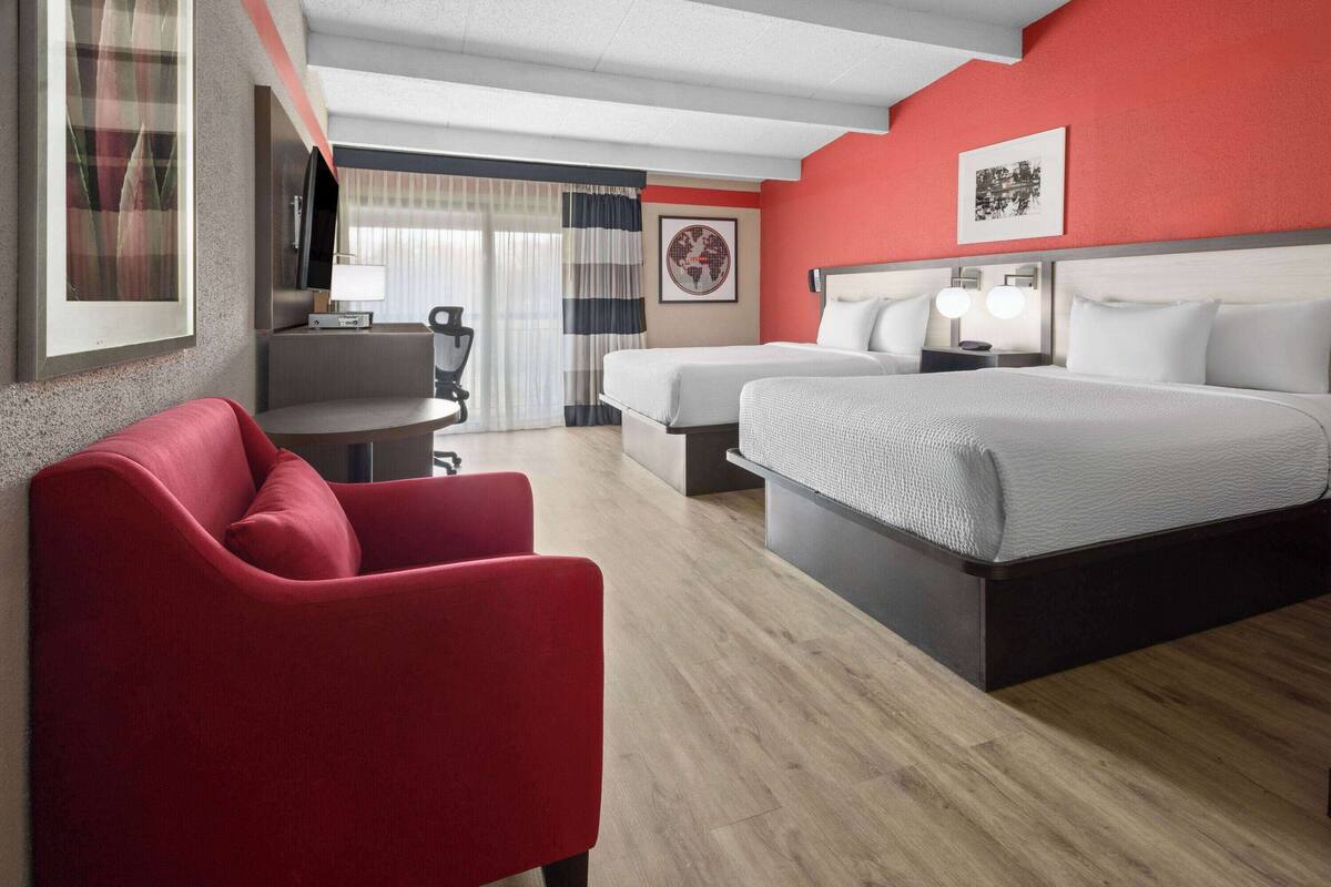 A hotel room with a red accent wall, two beds, a red armchair, a desk with a lamp, and a TV.