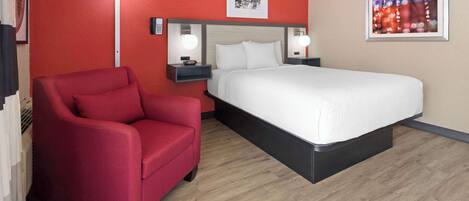 A hotel room with a red wall, a white bed, a red armchair, and a floor lamp.