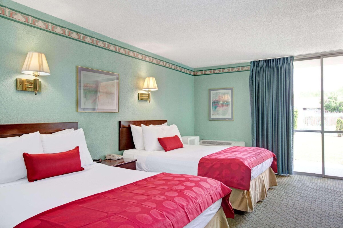 A hotel room with two beds, a large window with a view, and wall-mounted lamps.