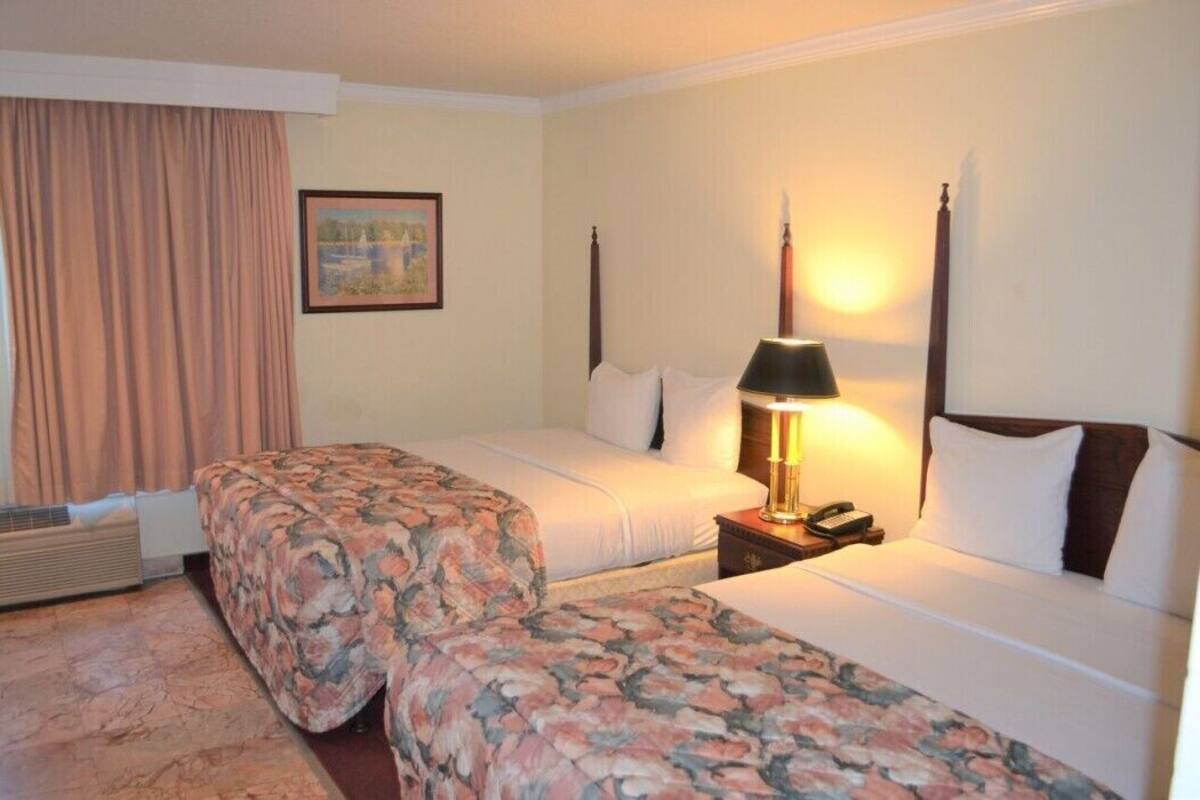 A hotel room with two beds, a nightstand with a lamp, a painting on the wall, and a window with curtains.