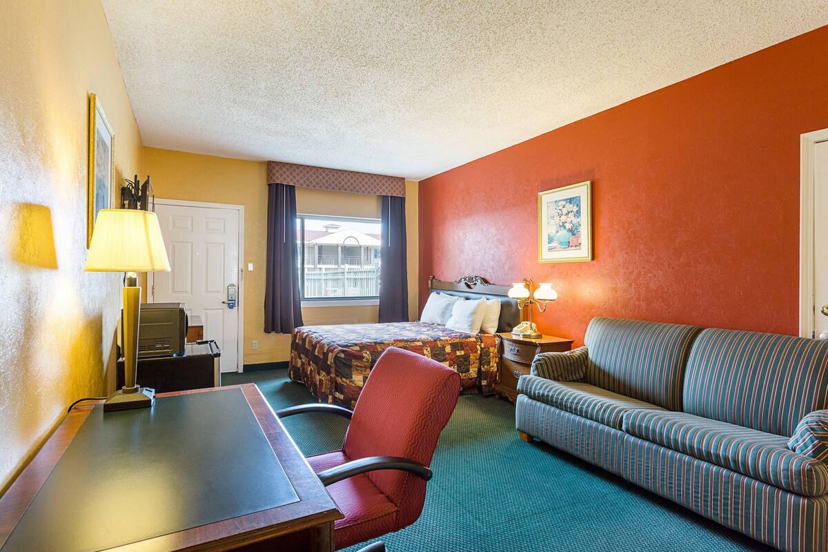 A hotel room with a bed, a desk, a chair, a sofa, a lamp, a nightstand, and a painting on the wall.