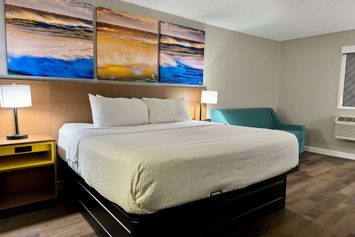 A hotel room with a large bed, two bedside lamps, a teal armchair, and three wall-mounted paintings of a sunset over the ocean.