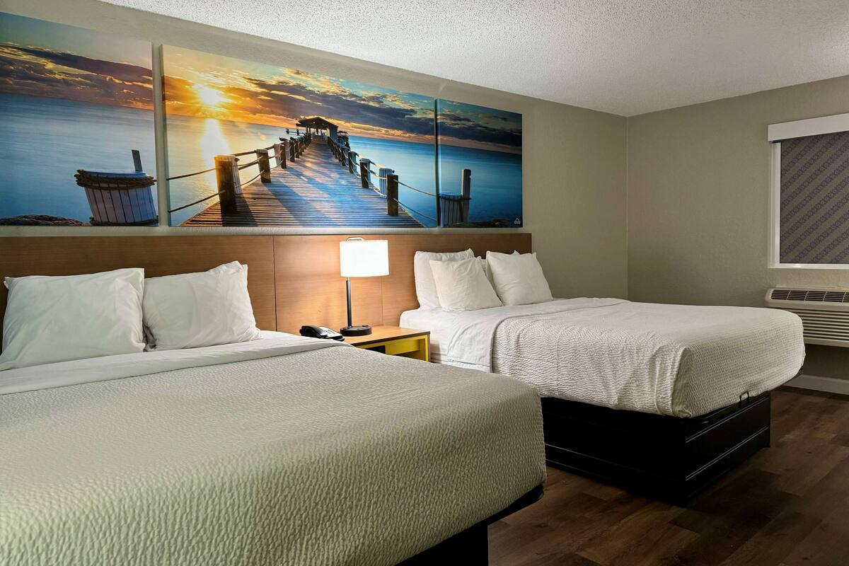 A hotel room with two beds, a nightstand, a lamp, and a large painting of a pier and sunset.