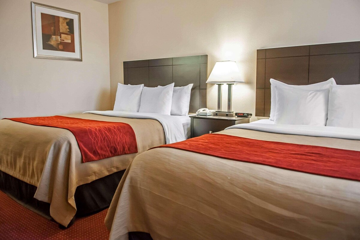 Habitación de hotel con dos camas, cada una con cabecera, mesita de noche con lámpara y un cuadro enmarcado en la pared.