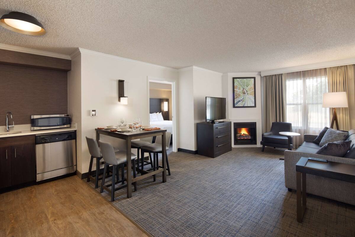 A modern hotel room with a kitchen, dining area, and living room. It includes a flat-screen TV, a fireplace, and a sofa.
