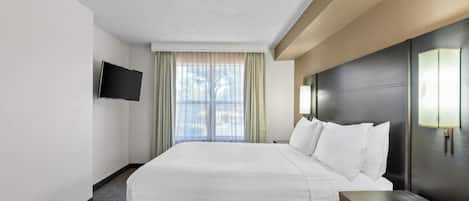 A hotel room with a bed, a flat-screen TV mounted on the wall, a window with curtains, and a nightstand with a lamp.