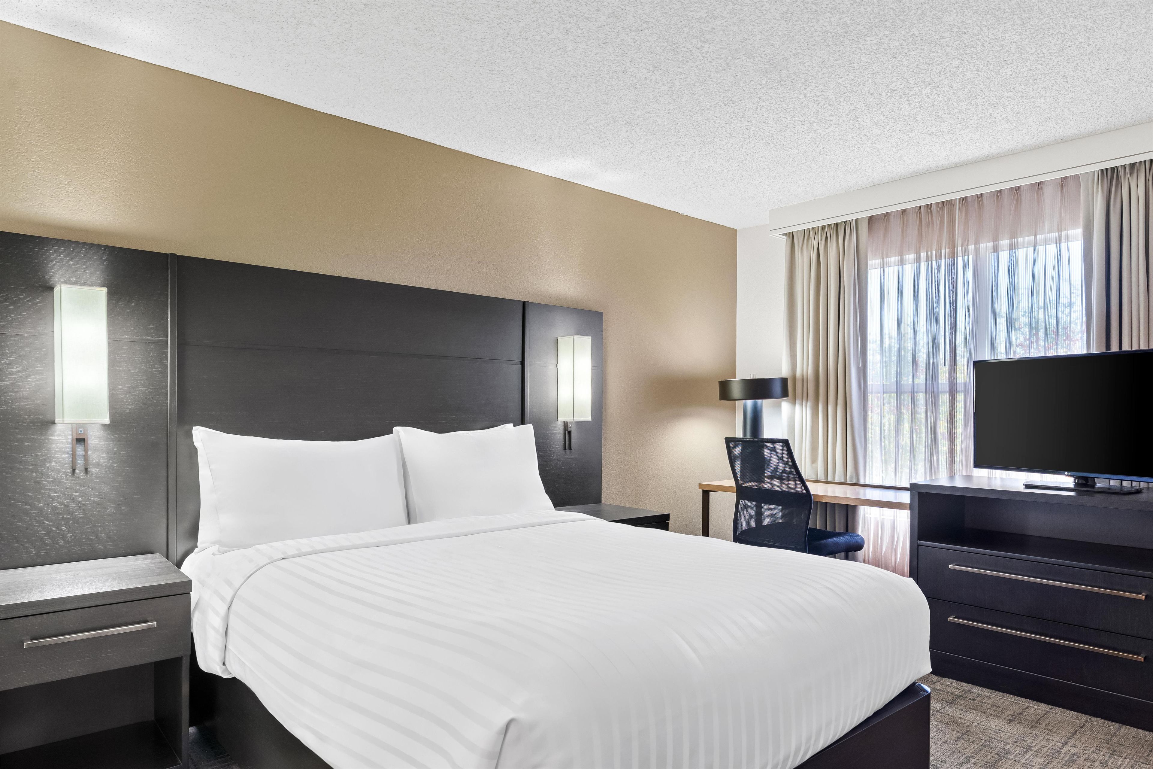 A hotel room with a bed, a desk, a chair, a TV, and a window with curtains.