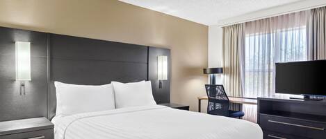 Una habitación de hotel con una cama, un escritorio, una silla, un televisor y un ventanal con cortinas.