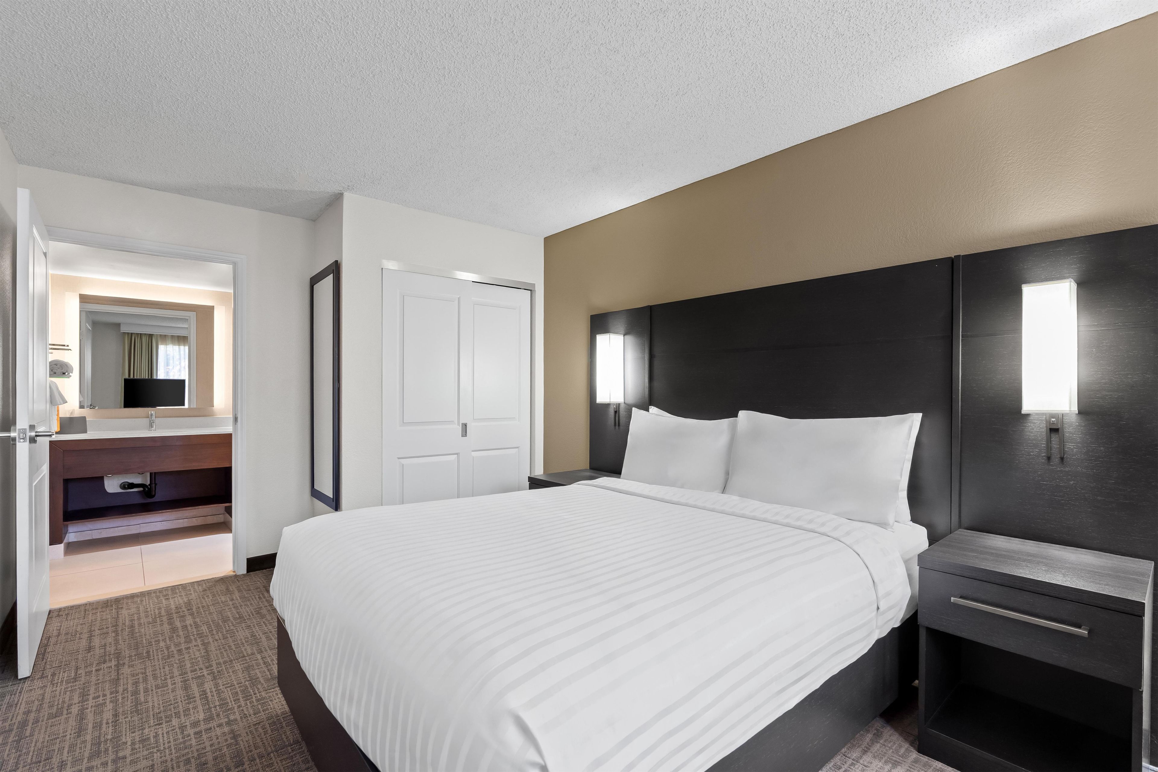 A hotel room with a bed, bedside table, nightstand, and a bathroom visible through an open door.