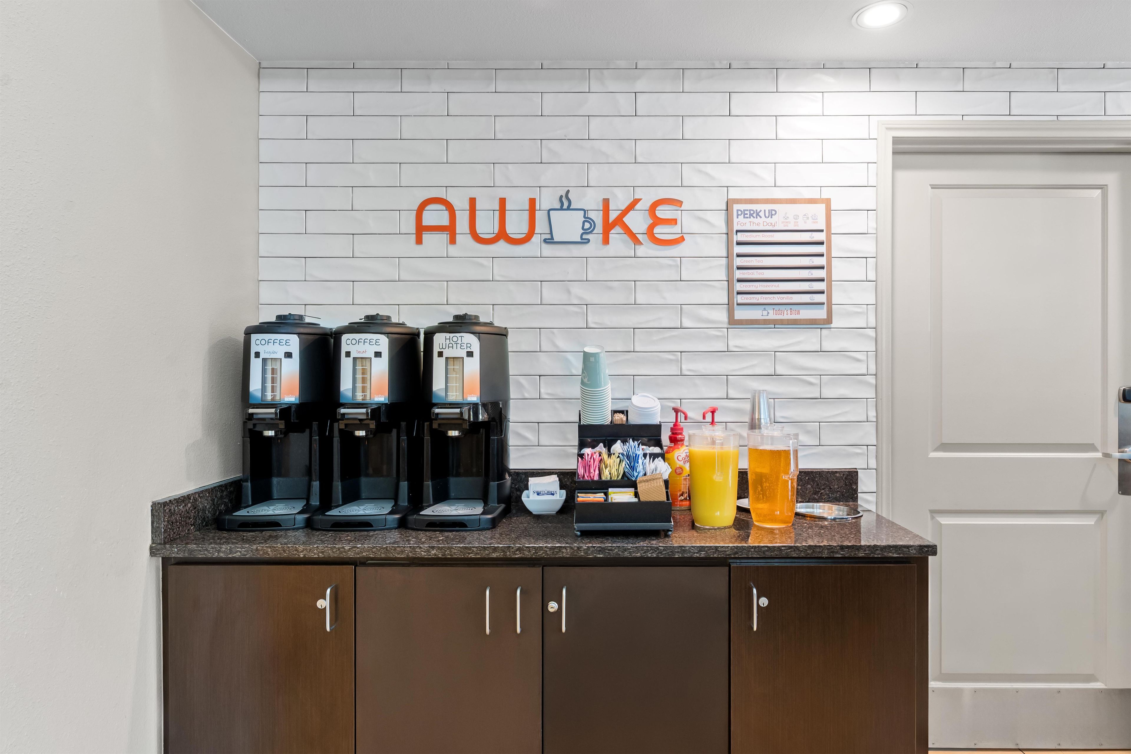 Un espace dédié au café avec plusieurs distributeurs, un panneau indiquant « AWOKE » et un tableau affichant le menu, ainsi qu’une porte sur la droite.