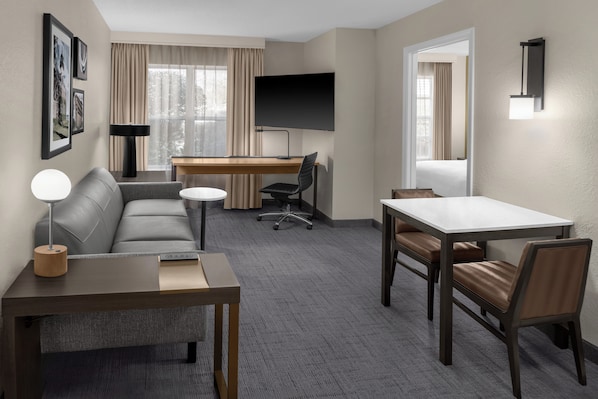 A hotel room with a sofa, a desk, a TV, a lamp, a table, and a chair. - Residence Inn by Marriott Mystic Groton (Mystic)