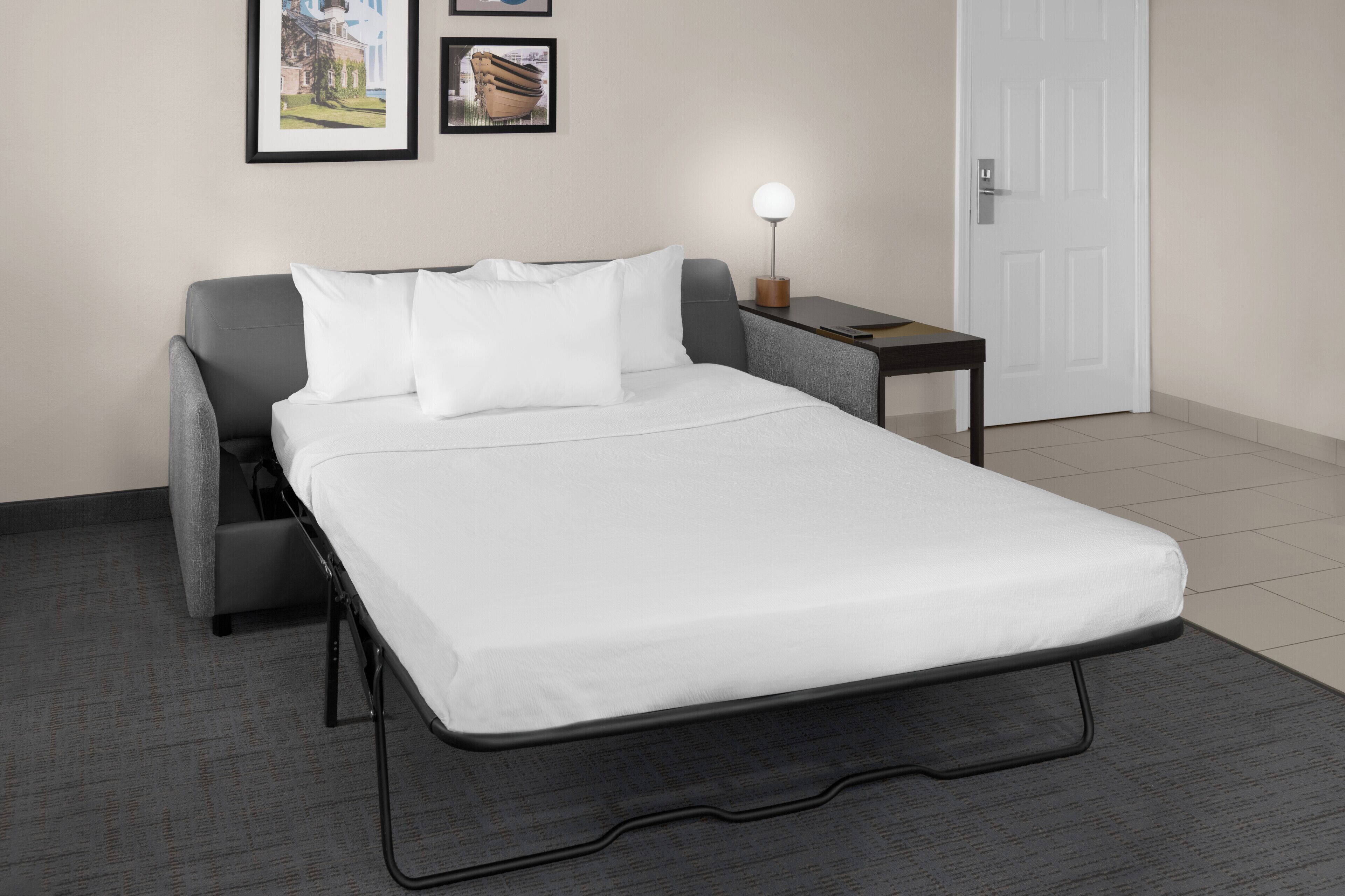 A single bed with a metal frame, grey upholstered headboard, and white bedding. A small bedside table with a lamp is to the right.