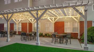 An outdoor patio area with tables and chairs, a pergola with string lights, and a brick building in the background.