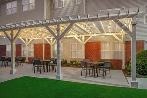 Un patio al aire libre con mesas y sillas, una pérgola con luces colgantes y un edificio de ladrillo al fondo.
