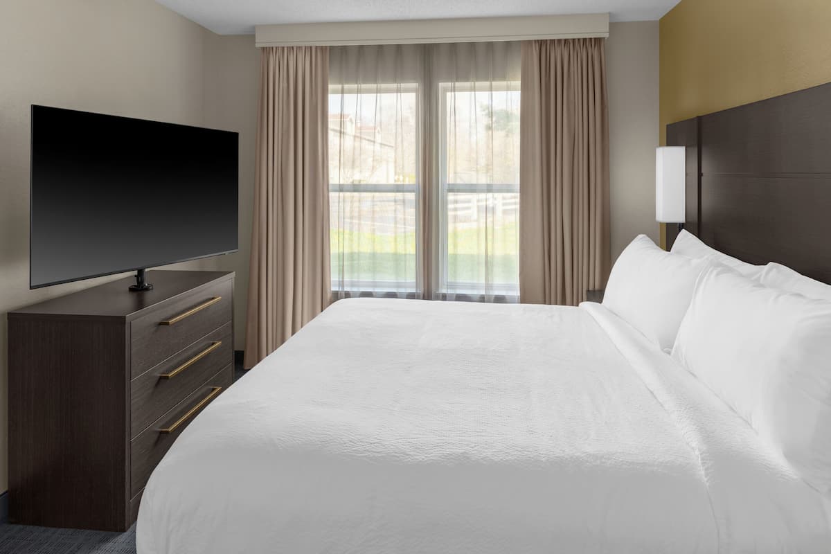 A hotel room with a large bed, a flat-screen TV, a dresser, and a window with curtains.