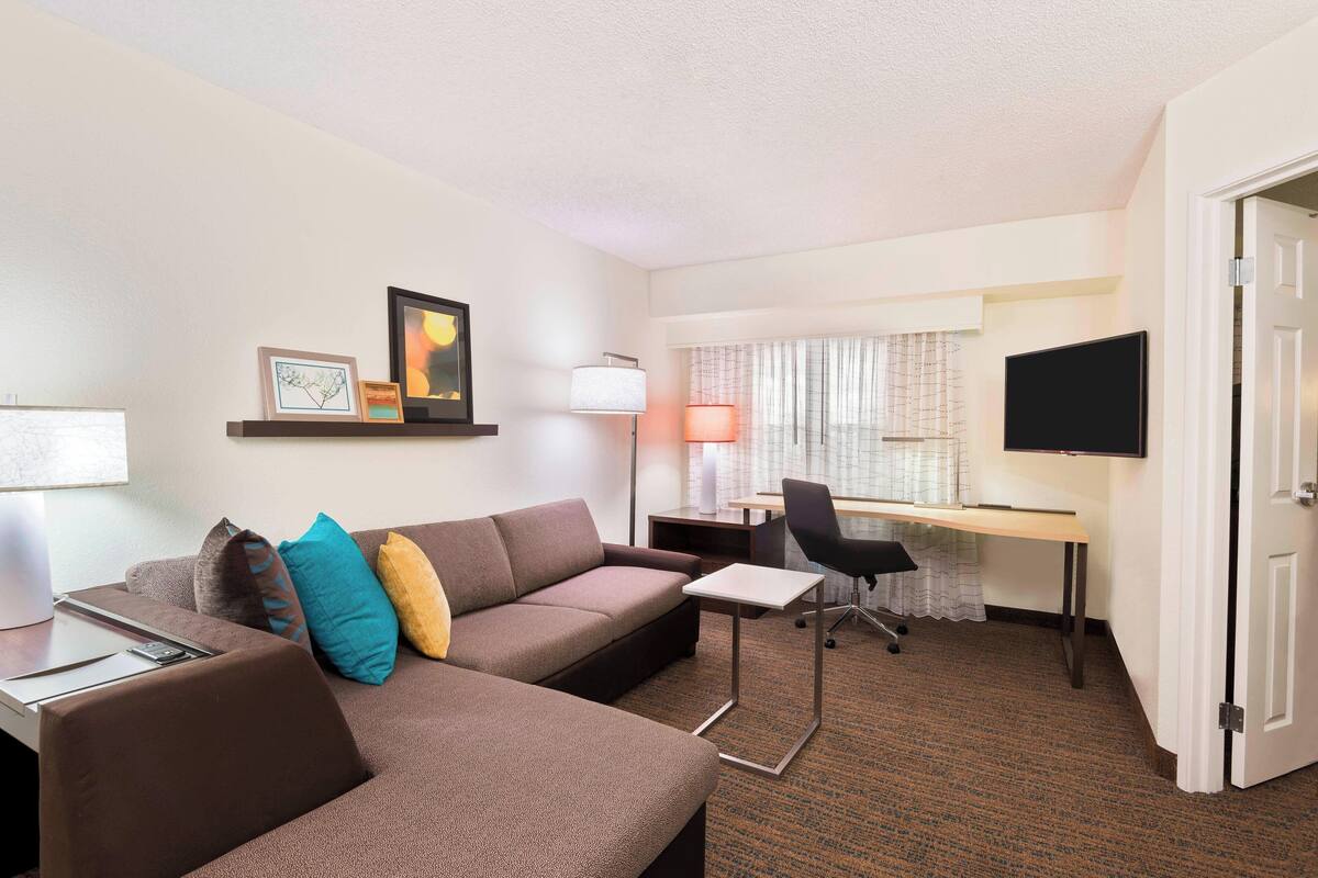 A hotel room with a sofa, a desk, a chair, a TV, and a lamp.