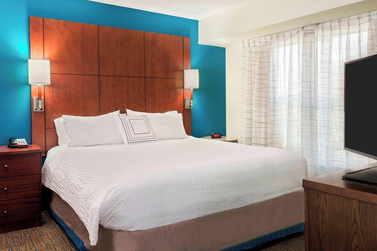 A hotel room with a large bed, wooden headboard, two bedside lamps, a TV, and a nightstand with a phone.