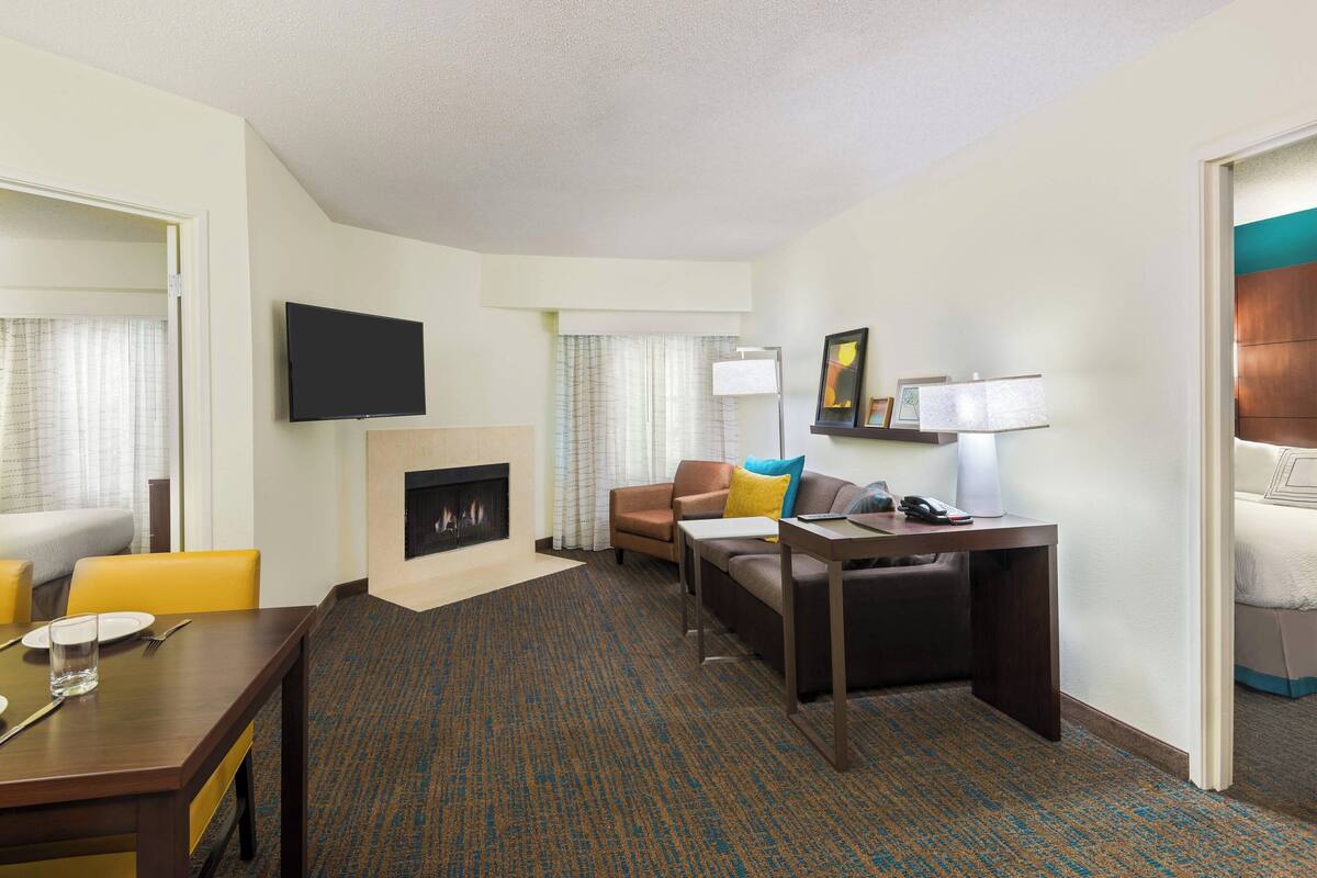 A hotel room with a living area featuring a sofa, armchairs, a dining table, a flat-screen TV, a fireplace, and a lamp.