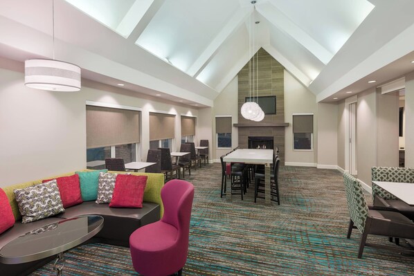 A modern hotel lobby with a high ceiling, seating areas, a fireplace, and a dining table.