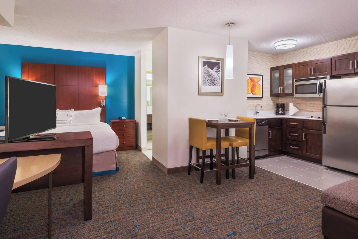 A hotel room with a bed, a desk with a computer, a chair, a small table with two chairs, a kitchen area with a microwave, a refrigerator, and a sink.