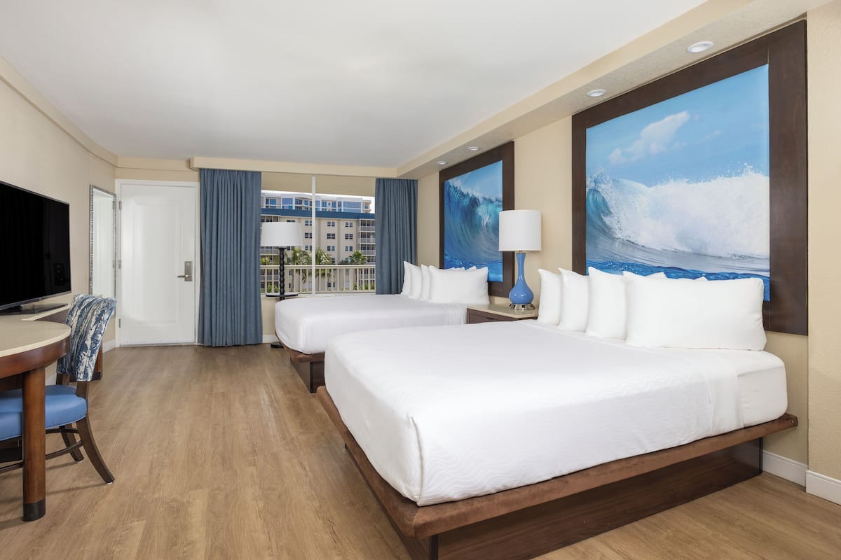 A hotel room with two beds, a TV, a desk, and a large window with a view of a building and palm trees.