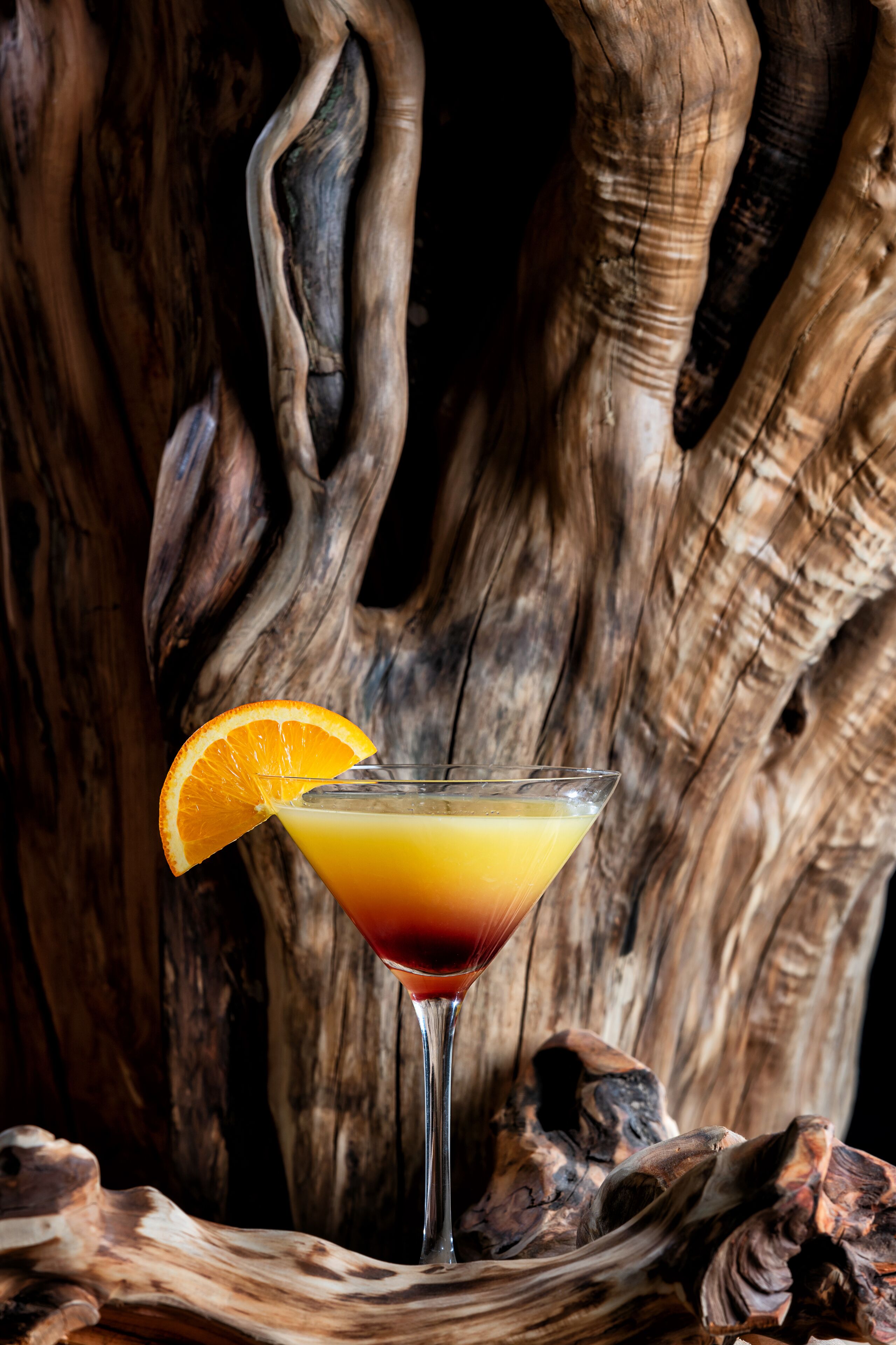 A cocktail glass with a slice of orange on the rim, placed on a wooden surface with a rough texture.