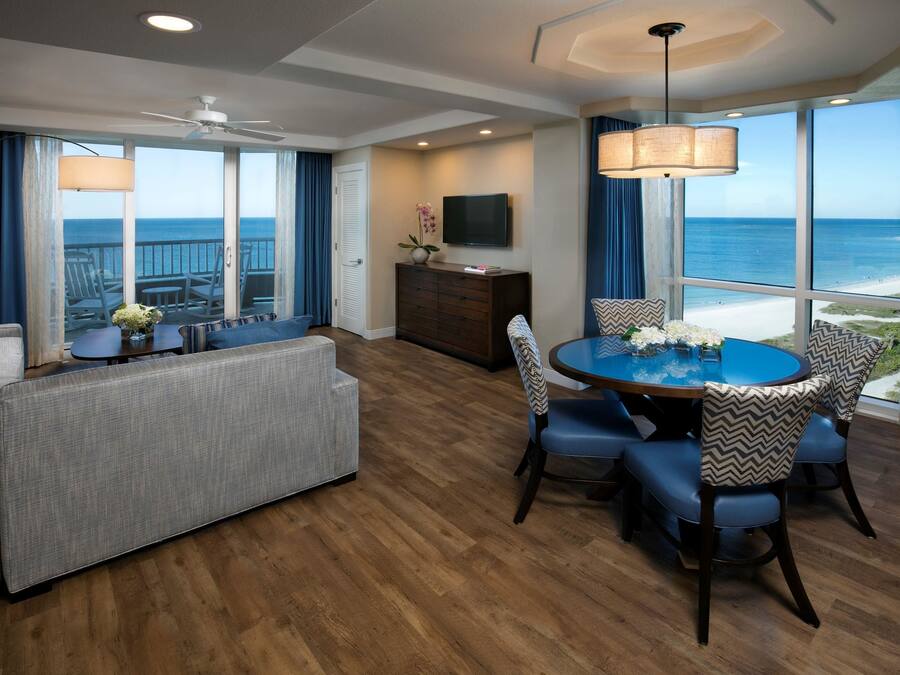 Een moderne hotelkamer met balkon, een zithoek met blauwe stoelen, een bank en een televisie aan de muur.