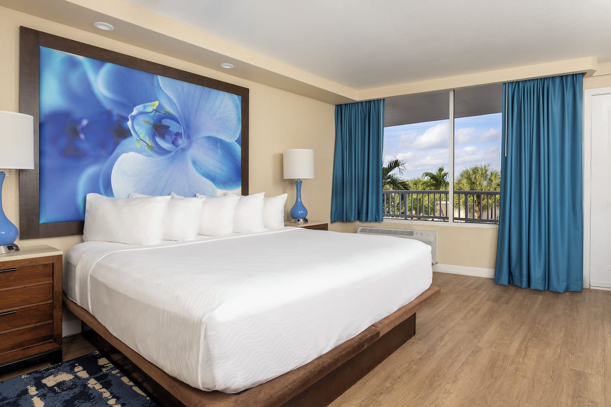 A hotel room with a large bed, a nightstand, a lamp, a large window with curtains, and a view of palm trees.