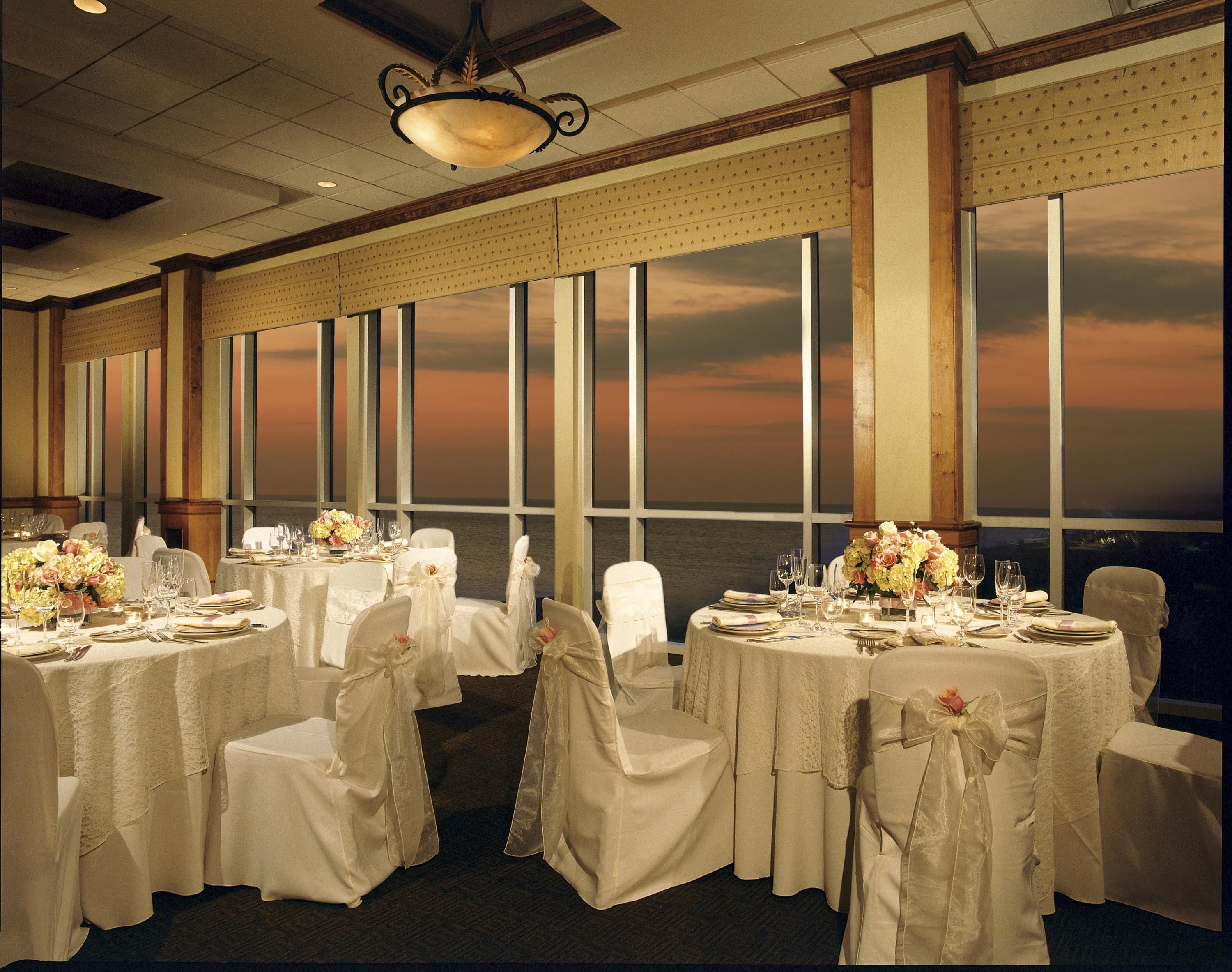 A banquet hall with round tables set for a formal event, large windows with a view of the sunset, and a classic chandelier.