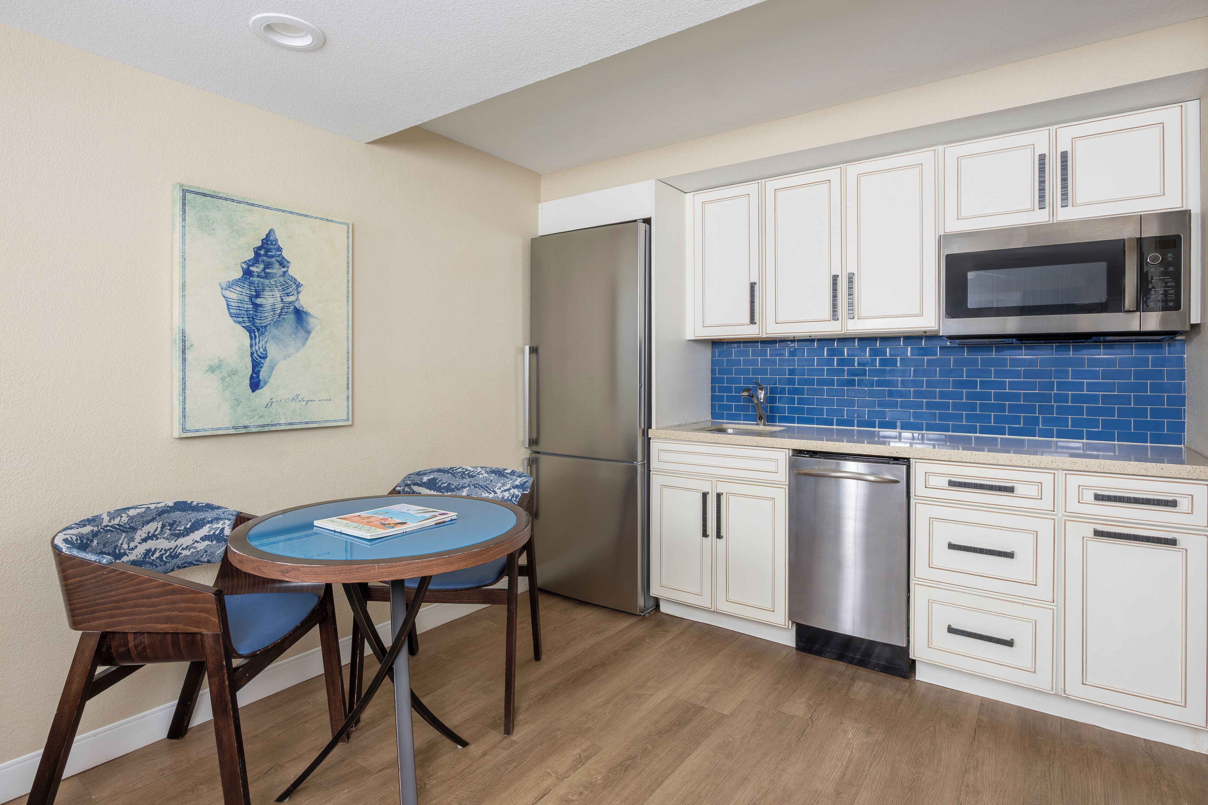A kitchen with white cabinets, a stainless steel refrigerator, a microwave, and a dishwasher. There is a blue tiled backsplash and a framed picture on the wall.