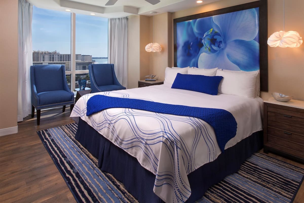 A hotel room with a large bed, a ceiling fan, a view of the city, a blue chair, and a large window.