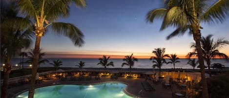 Un espace piscine avec des chaises longues et des palmiers, offrant une vue sur l’océan au coucher du soleil.