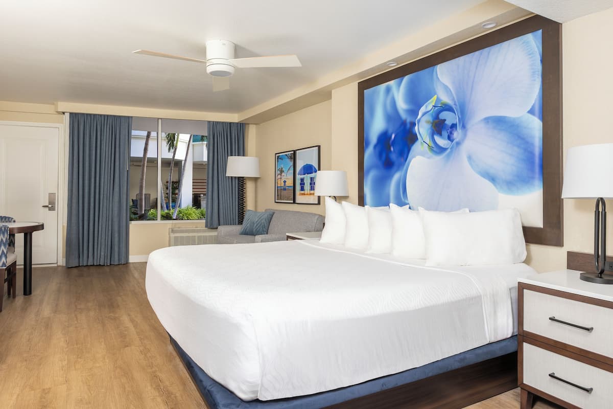 A hotel room with a large bed, a desk, a chair, a ceiling fan, and a large window with curtains.