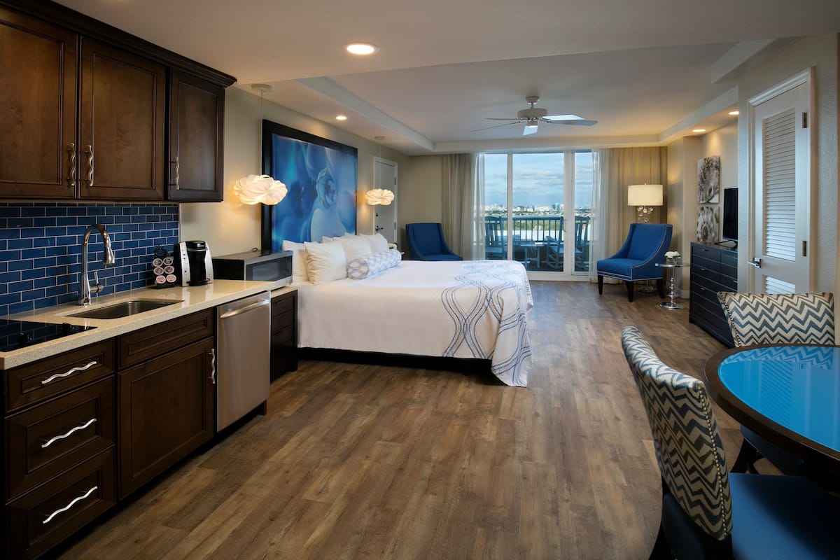 A hotel room with a large bed, a dining area with a table and chairs, a kitchen with a sink and cabinets, and a balcony with a view.