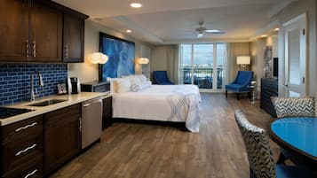 A hotel room with a large bed, a dining area with a table and chairs, a kitchen with a sink and cabinets, and a balcony with a view.
